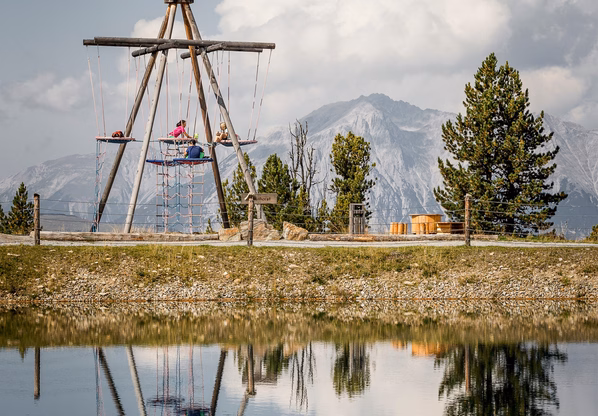 Abenteuerspielplatz mit Spiegelung im Bergsee Ein Holzklettergerüst auf dem Spielplatz spiegelt sich im ruhigen Wasser, dahinter stehen Bäume und Berge.