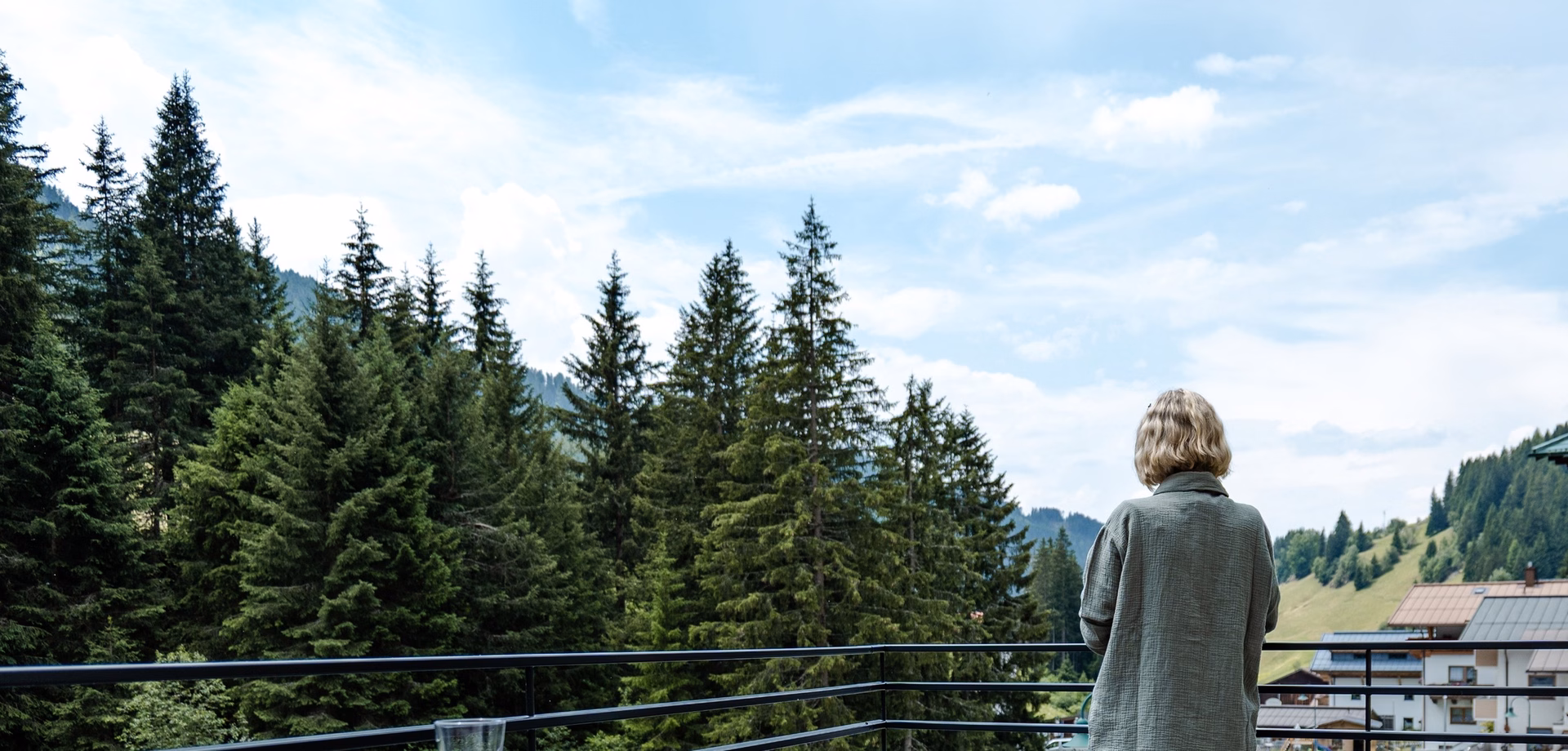 A person stands on the balcony looking out over the forest and mountains.