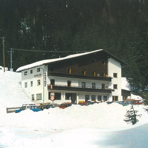 Ein verschneites Haus mit der Beschriftung PENSION und GASTHOF steht am Hang, davor sind geparkte Autos zu sehen.