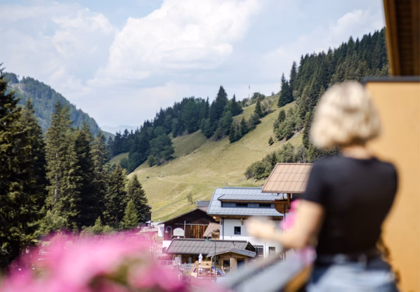 Bergblick vom Hotel Waldesruh in Ochsengarten Im Vordergrund steht eine unscharf dargestellte Person auf dem Balkon, dahinter sind Wiesen, Nadelwald und Häuser in den Bergen zu sehen.
