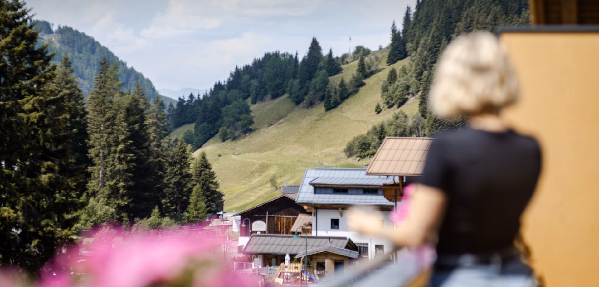 A view across the valley toward Hotel Waldesruh in the mountains, with blurred pink flowers and a person in the foreground.